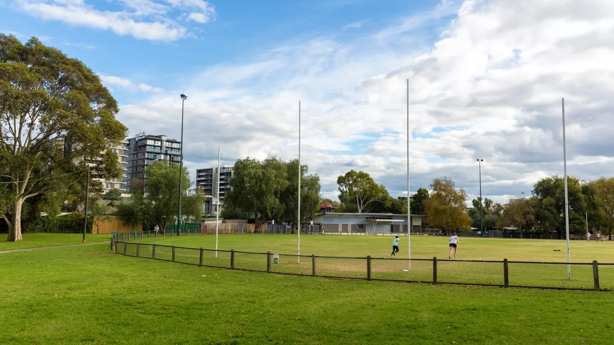 A grassy park with two people playing sports, surrounded by trees and modern buildings under a partly cloudy sky.