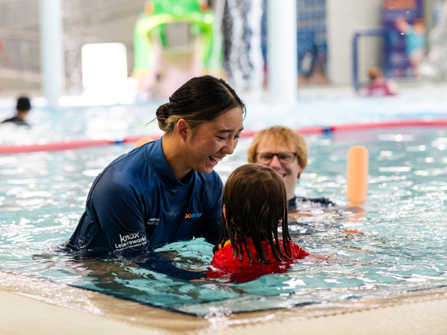 a child in a red swimming top with 2 swim teachers in the pool.