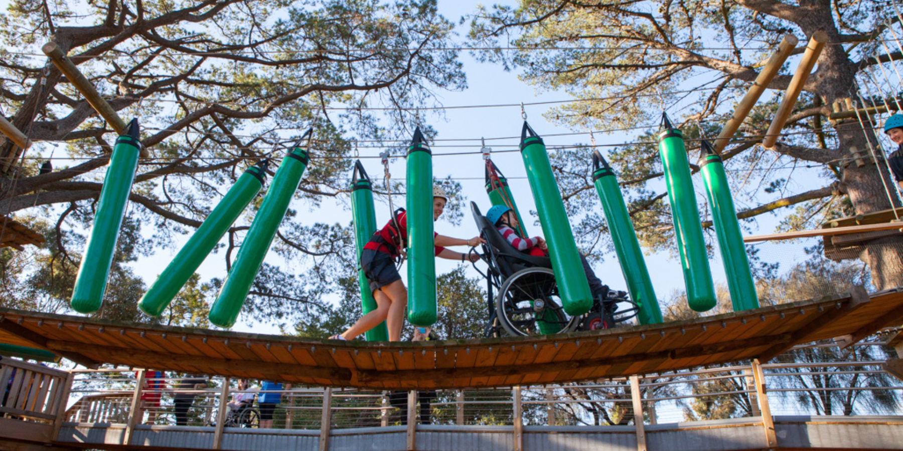 A wheelchair user is harness and being pushed along a ropes course