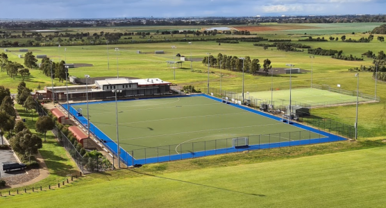 Aerial view of a sports complex featuring a blue hockey field, a clubhouse, and surrounding green fields under a cloudy sky.
