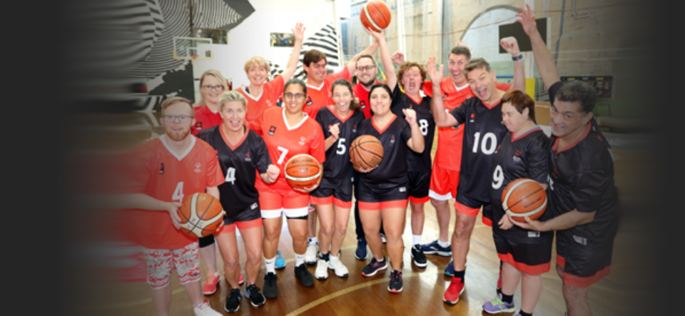 A diverse group of enthusiastic basketball players celebrate together, wearing team jerseys and holding basketballs in a gym.