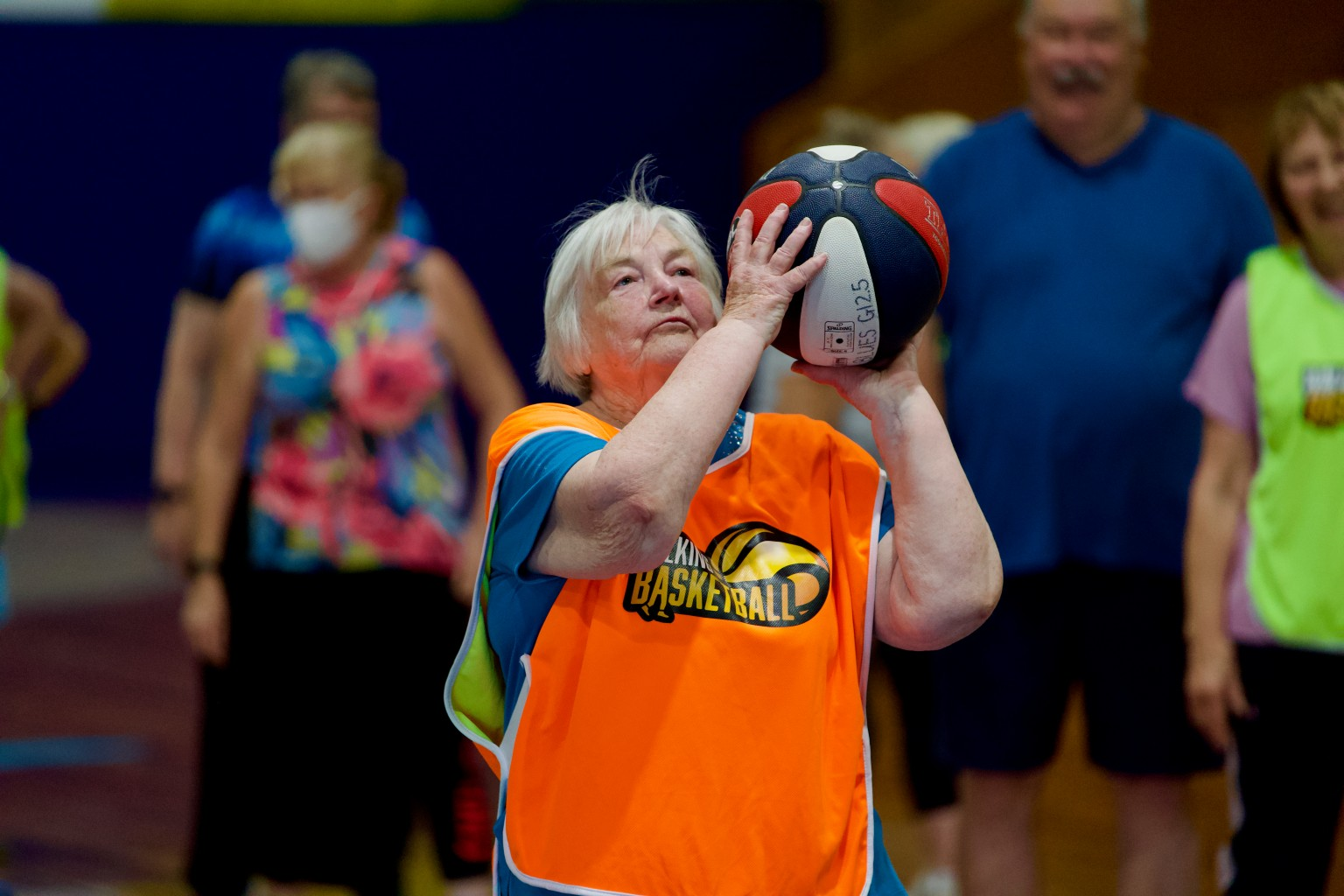 A woman wearing an orange basketball jersey holds a basketball, preparing to make a shot in a sports gym with others watching.
